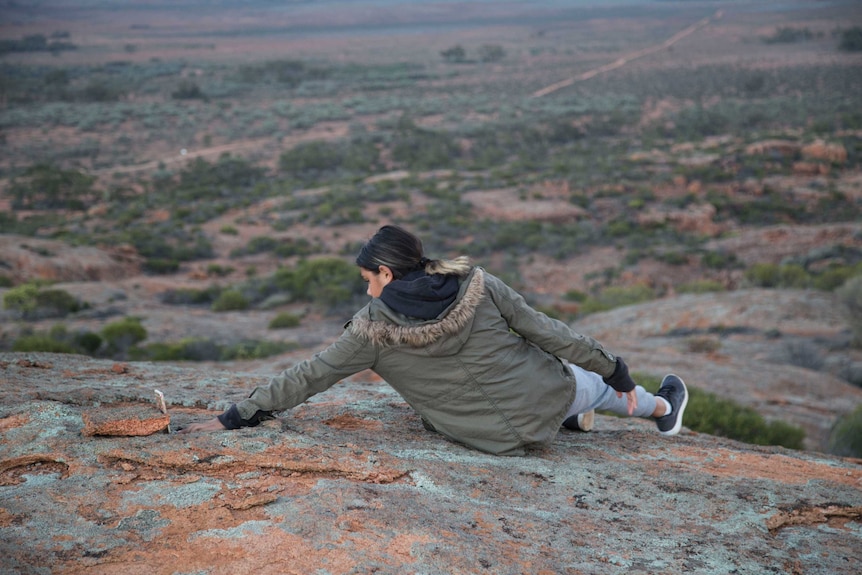 A young woman reaches for her mobile phone to take a photo while lying across a rock.