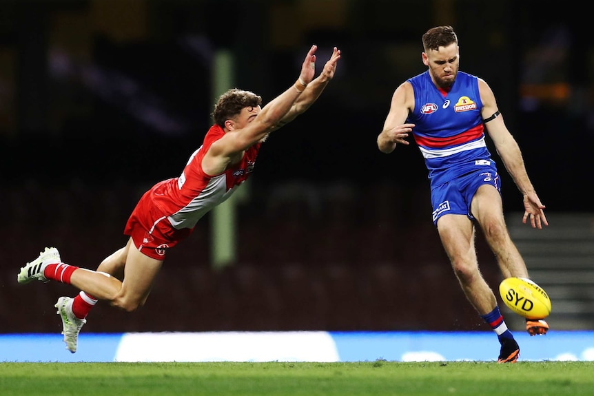 A Western Bulldogs AFL player kicks the ball with his left foot as a Sydney Swans opponents dives in the air to smother.