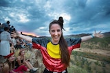 woman stands with arms extended out, wearing sequined tshirt of Aboriginal flag, in front of festival goers and sunset.