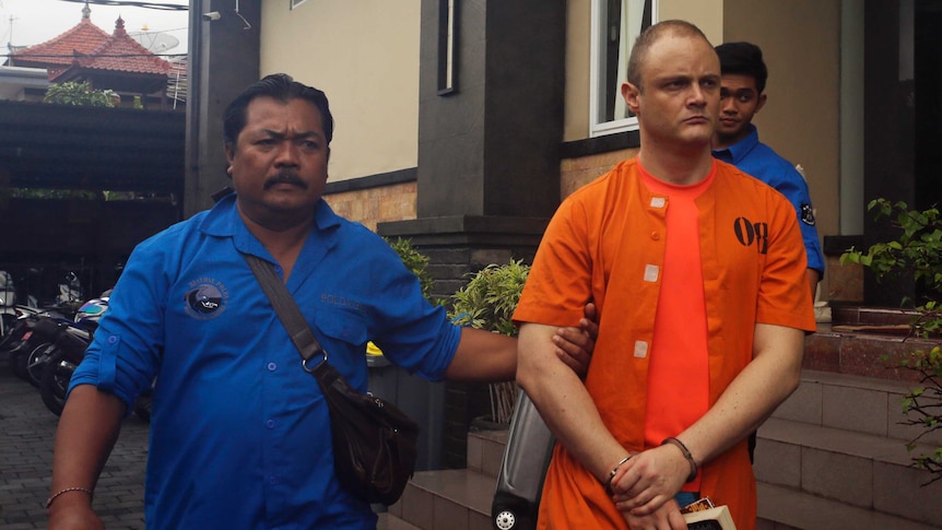 Isaac Emmanuel Roberts wears an orange jumpsuit and is escorted from a building in Bali.