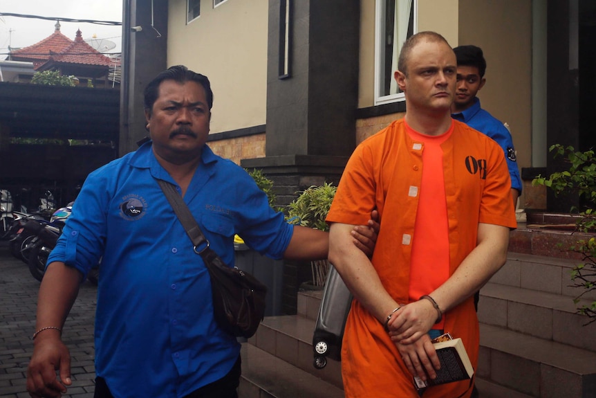 Isaac Emmanuel Roberts wears an orange jumpsuit and is escorted from a building in Bali.