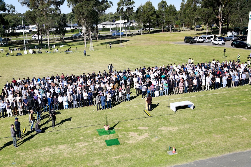 Birds-eye view of a large group of mourners on grass sitting in rows with a casket at the front.