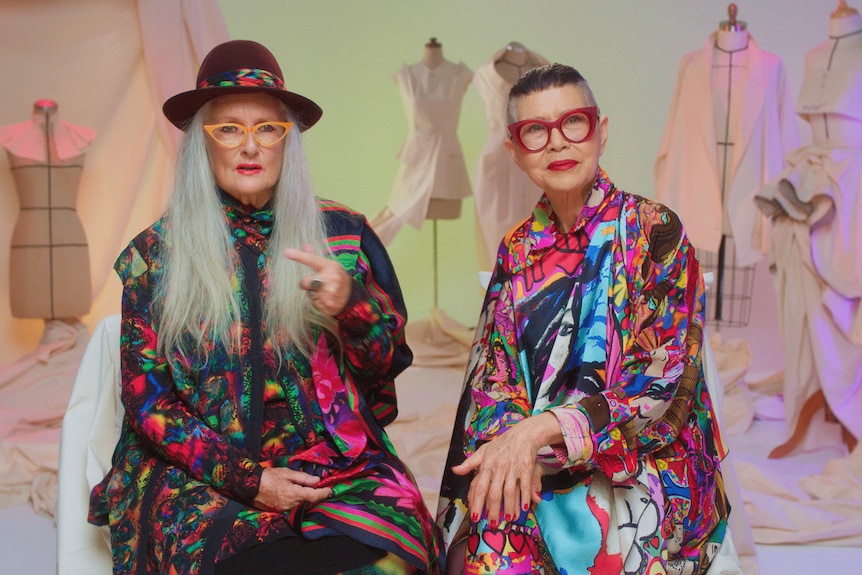 Linda Jackson and Jenny Kee, wearing bright rainbow coloured clothes, sitting in front of a tv set with fashion models behind
