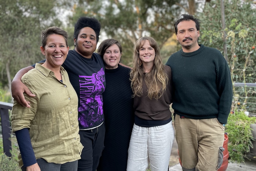 Five people arm in arm on a garden deck in the countryside, smiling. 