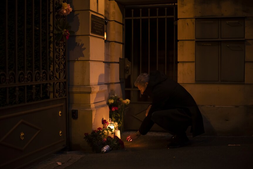 A man kneels to light a candle in front of a stone gatepost, in darkness.