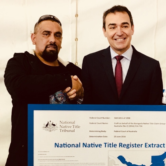 Picture of two men next to each other, shaking hands in front of a big poster with "national native title register" on it