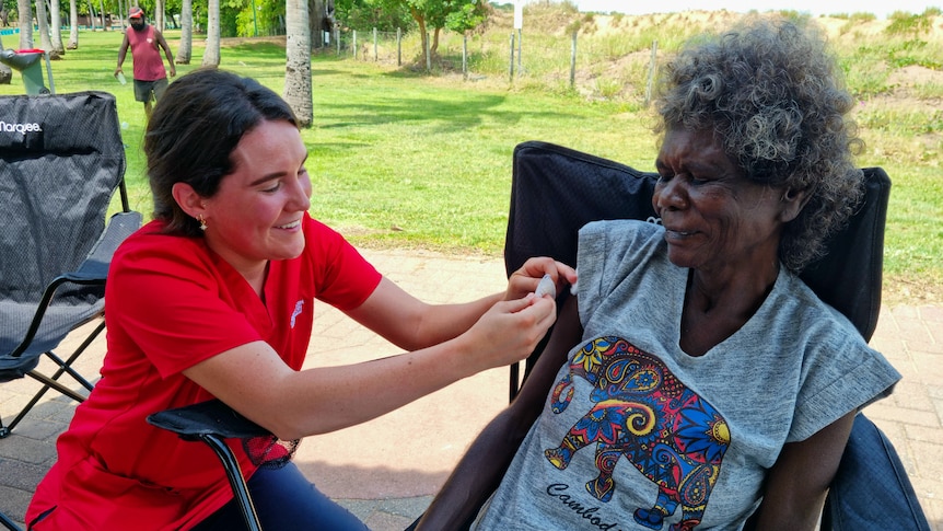 A woman applying a bandaid to another woman's arm after vaccinating her, with greenery in the background.