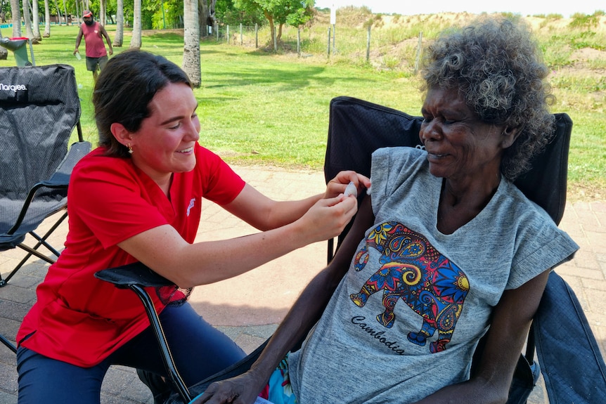 A woman applying a bandaid to another woman's arm after vaccinating her at a park, with greenery in the background.