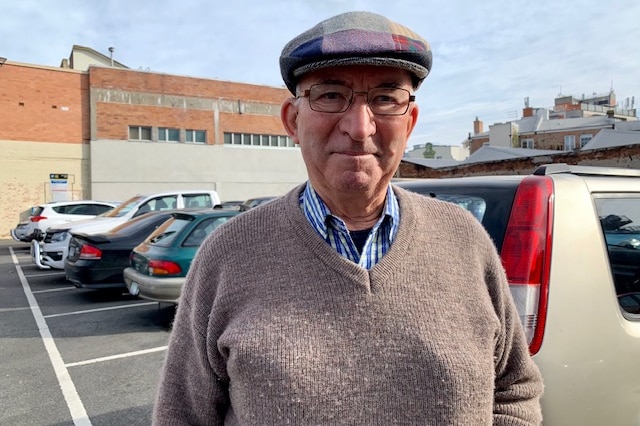 An elderly man wearing glasses and a checked cap stands in a carpark.