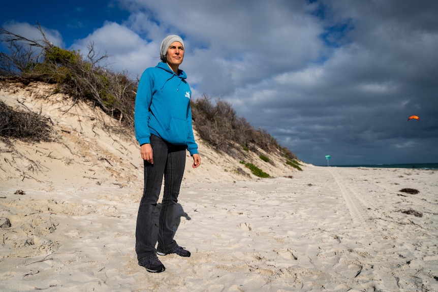 A woman stands on a beach wearing black pants, a blue jumper and a beanie, looking out towards the water.
