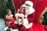A toddler cries with his arms in the air while sitting on a lap of a man dressed in  red Santa costume.