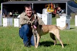 A man and his dog pose for a photograph in front of a row of honey bee hives.
