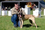 A man and his dog pose for a photograph in front of a row of honey bee hives.