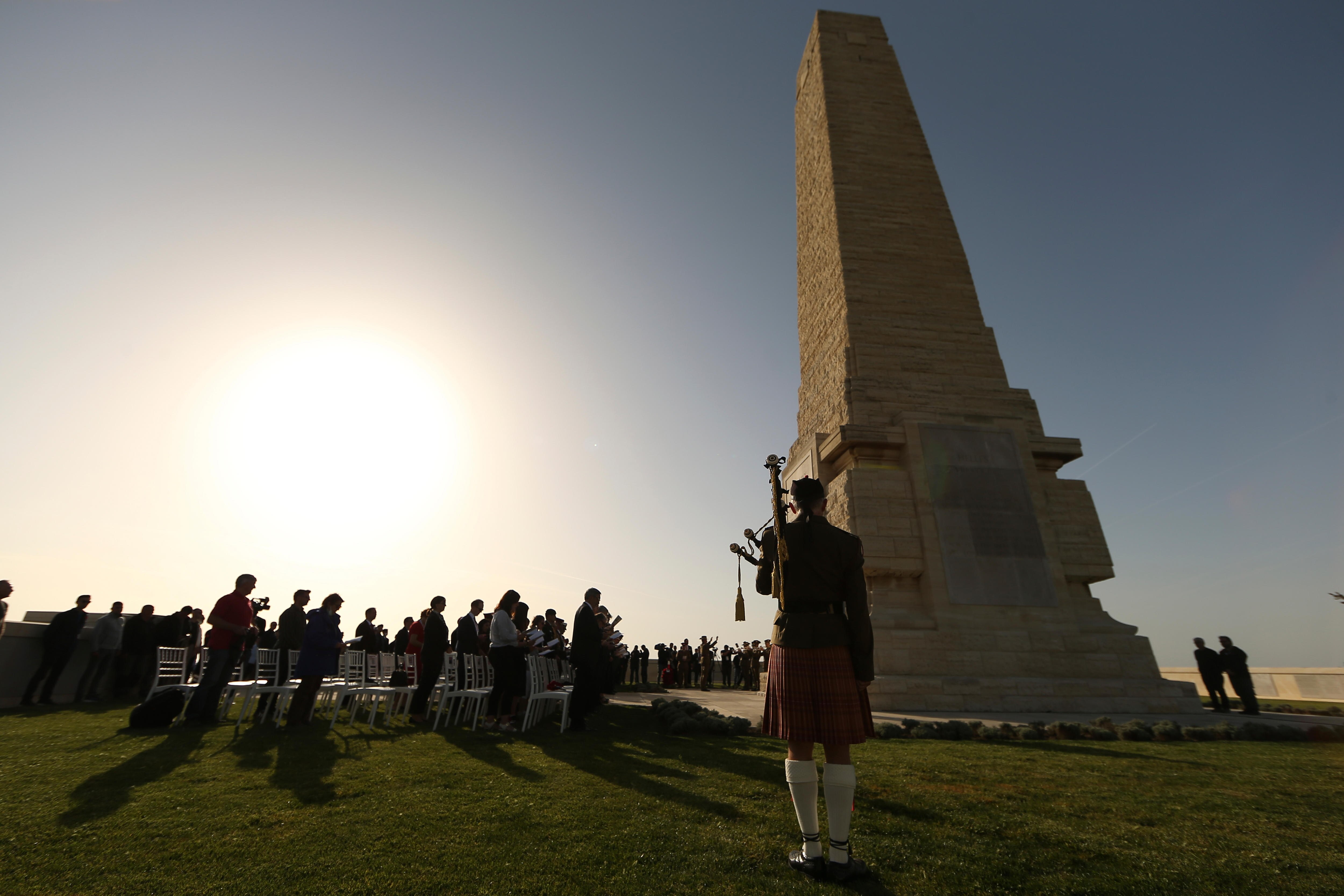 A figure dressed in Scottish garb holds bagpipes before a crowd with heads bowed and a cenotaph 