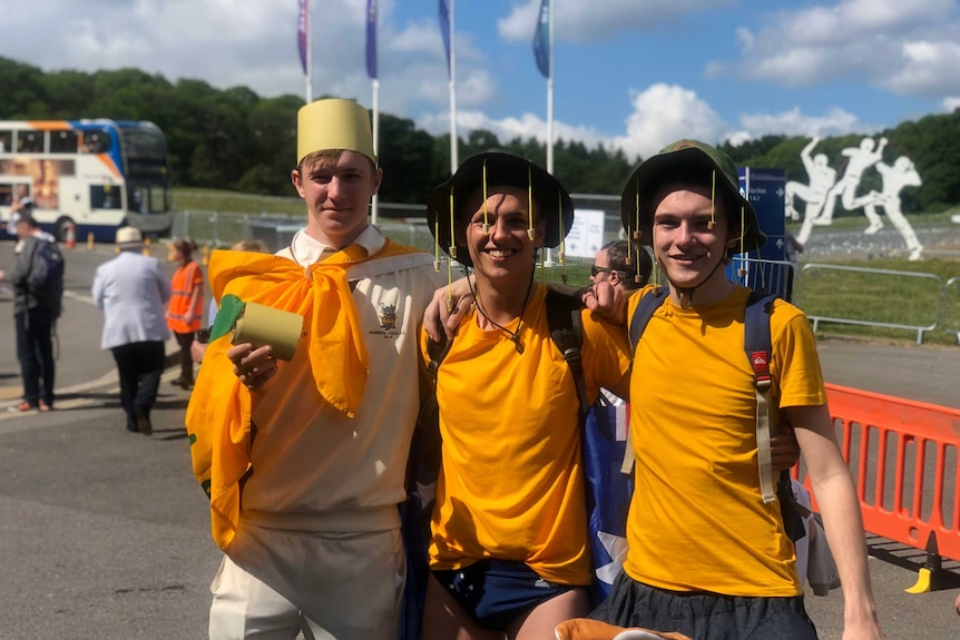 Two men wear gold t-shirts while another wears a cricket uniform with a green and gold flag tied around his neck