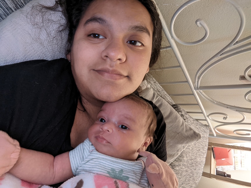 A young woman lays on a bed with her baby on her chest. They both look tired but happy
