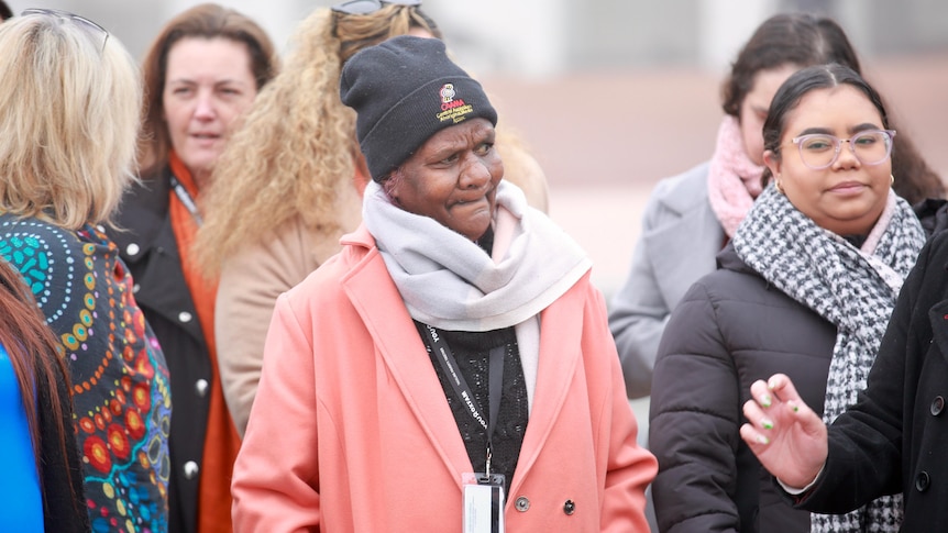 An indigenous woman in a pink coat, scarf and black beanie stands in middle of a group of women. 
