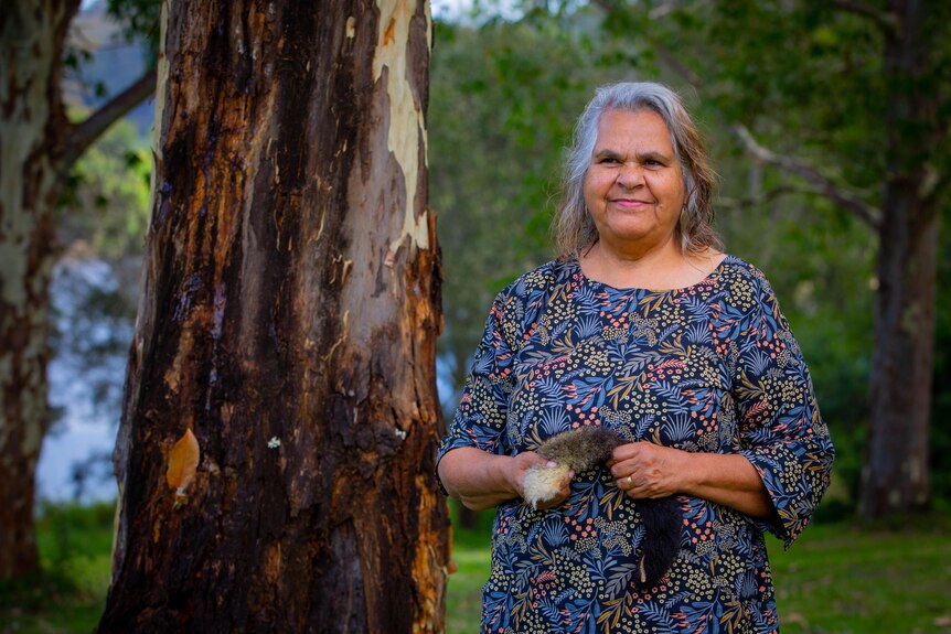 Aunty Loretta standing outdoors among bushland, holding a possum tail and smiling.