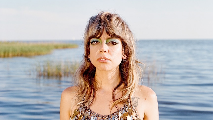 Alynda Segarra stands on the banks of a body of water. They wear eye makeup and a top made entirely out of keys.