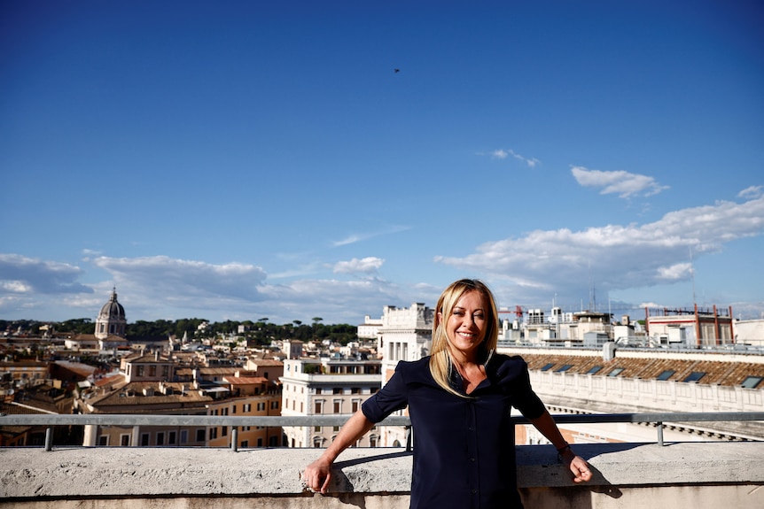 A woman with blonde hair poses against a wall with a backdrop of a city behind her.