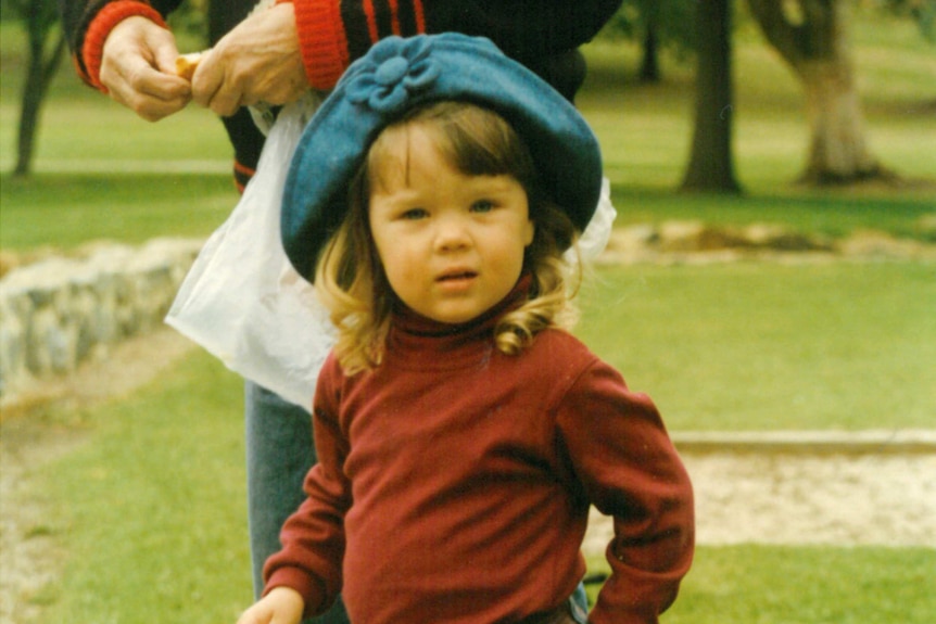 A toddler girl wearing a blue hat and jeans feeds ducks in a park.