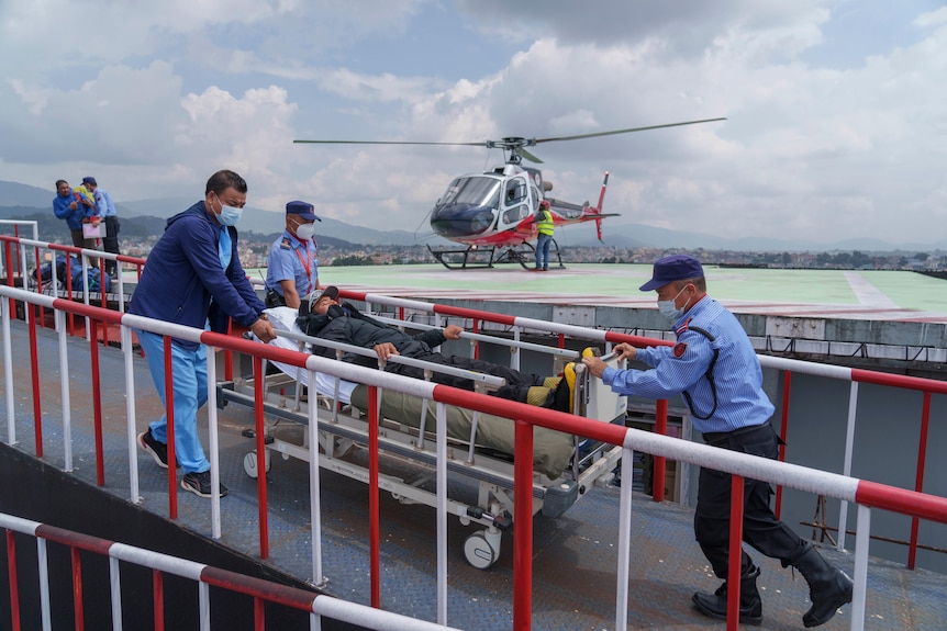 A young Nepalese man in winter clothing is wheeled on a hospital bed by health workers away from a helicopter landing pad.