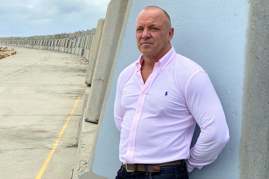 A man in a pink business shirt stands against a rockwall, serious facial expression