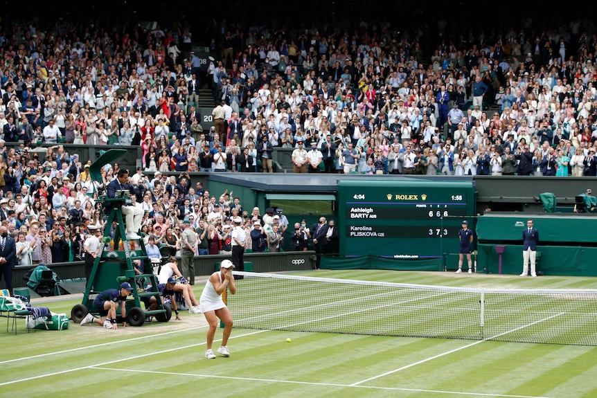 A young female tennis player in whites reacts with joy on a grass court in front of a packed stadium.