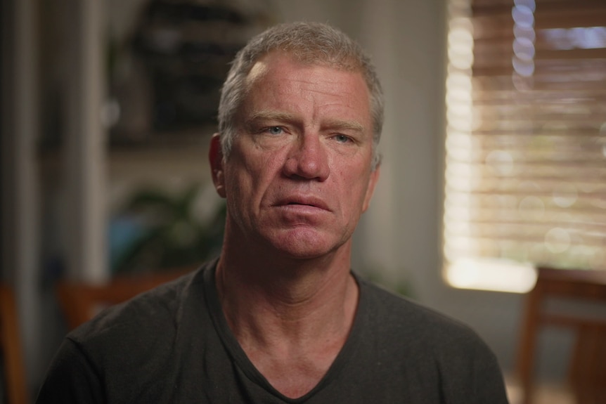 An older man dressed in a grey t-shirt looks seriously at the camera.