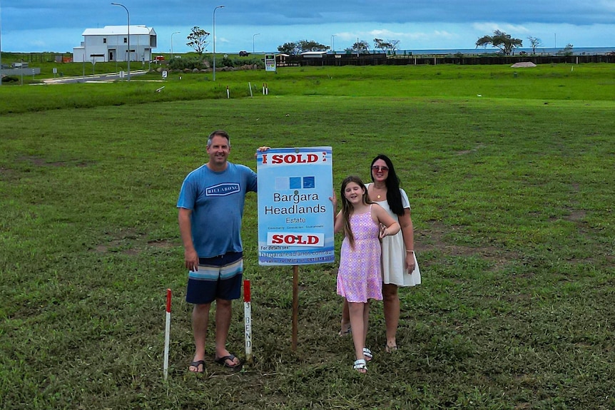 A man and his wife and daughter stand on a block of land next to a sold sign.