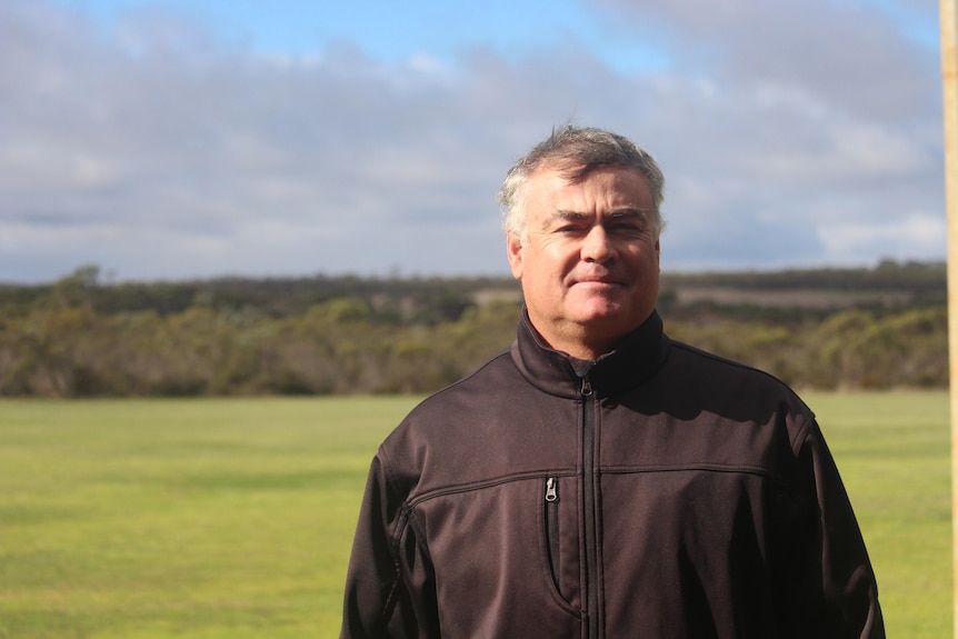 A grey-haired man wearing a brown winter jacket stands in a field, with a line of trees and a cloudy sky in the background
