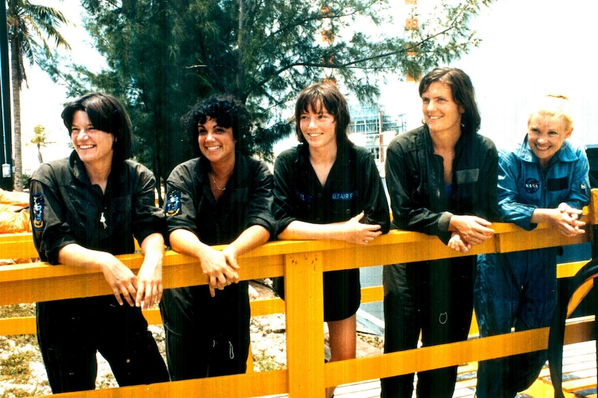 Five women smiling and leaning against a yellow fence