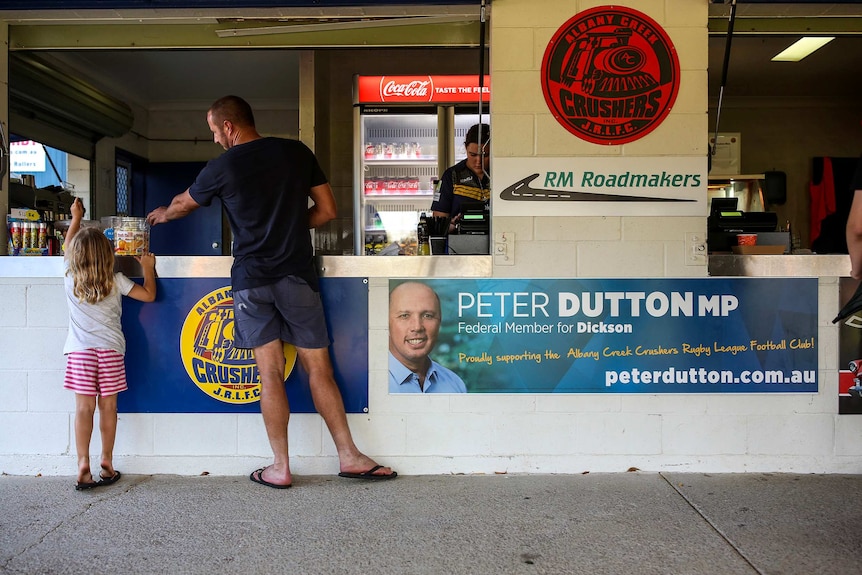 A man and girl stand at the window of a canteen at a junior rugby league football club next to a Peter Dutton sign.