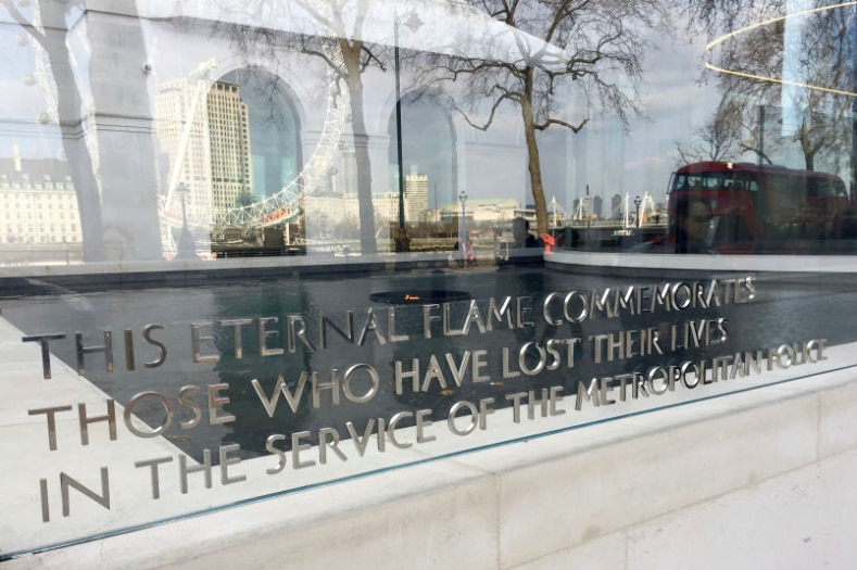 The eternal flame plaque at New Scotland Yard with the London Eye and a red double-decker bus reflected in it.