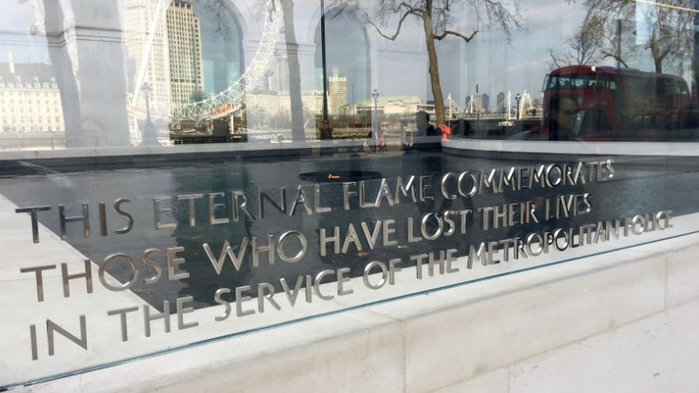 The eternal flame plaque at New Scotland Yard with the London Eye and a red double-decker bus reflected in it.
