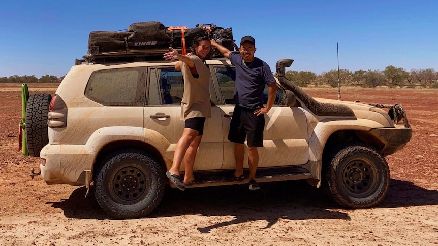 A man and a woman hang off the side of a mud-covered 4WD on an outback track.