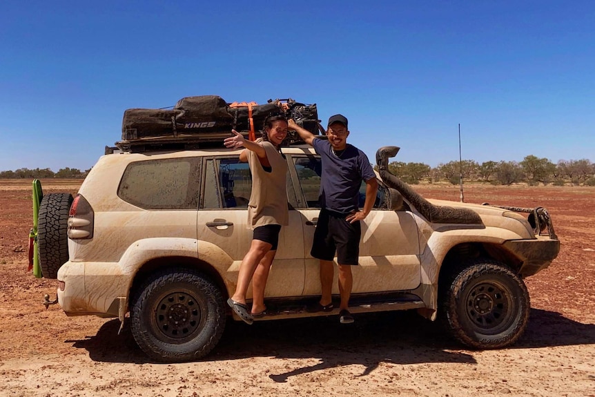 A man and a woman hang off the side of a mud-covered 4WD on an outback track.
