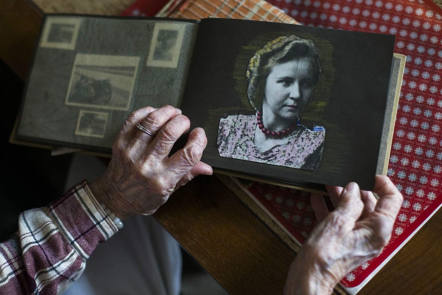 Two elderly hands hold a black and white photo album, the open page showing a woman in 1930s style hair and clothes.