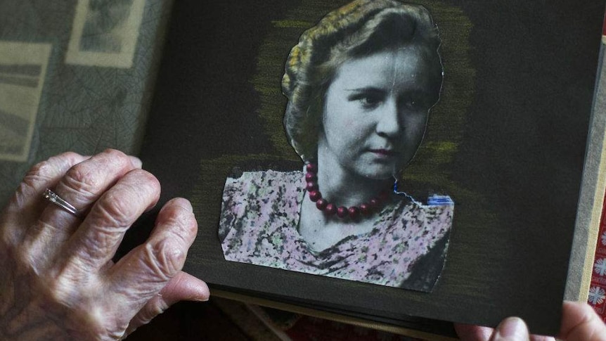 Two elderly hands hold a black and white photo album, the open page showing a woman in 1930s style hair and clothes.