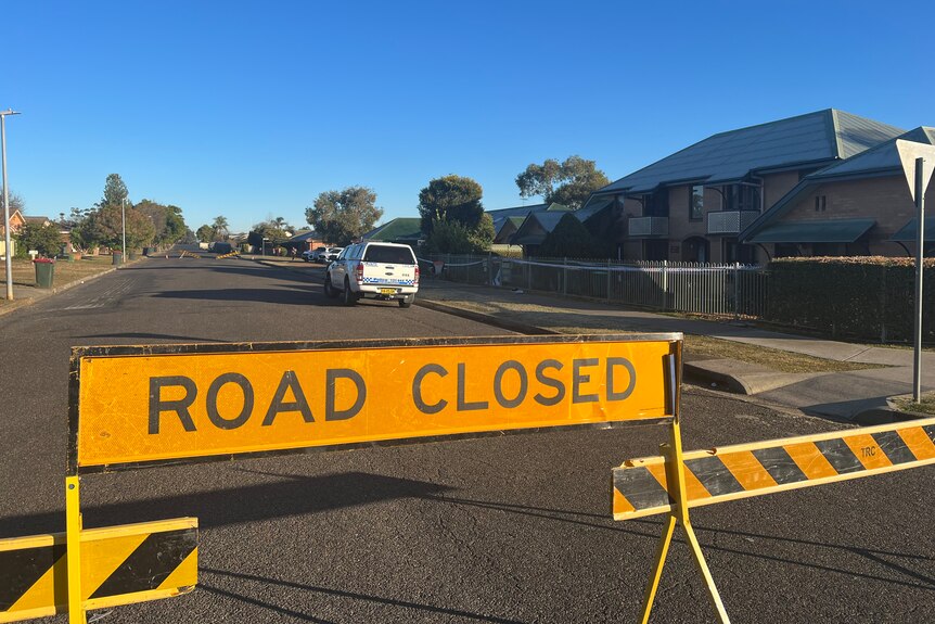 A road closed sign sits on s residential street behind it is a police car, police tape and a crime scene. 