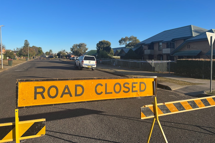 A road closed sign sits on s residential street behind it is a police car, police tape and a crime scene. 