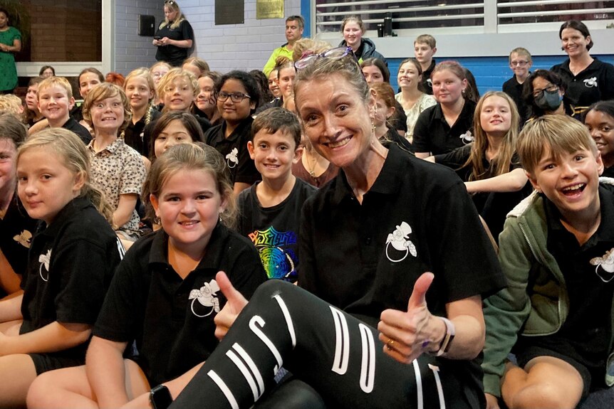 A smiling woman gives a thumbs up to the camera. She is sitting with a group of children, dressed in matching black shirts. 