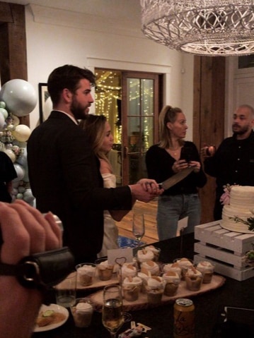 A man and a woman stand closely as both hold a large knife in their right hands and look towards a white cake.