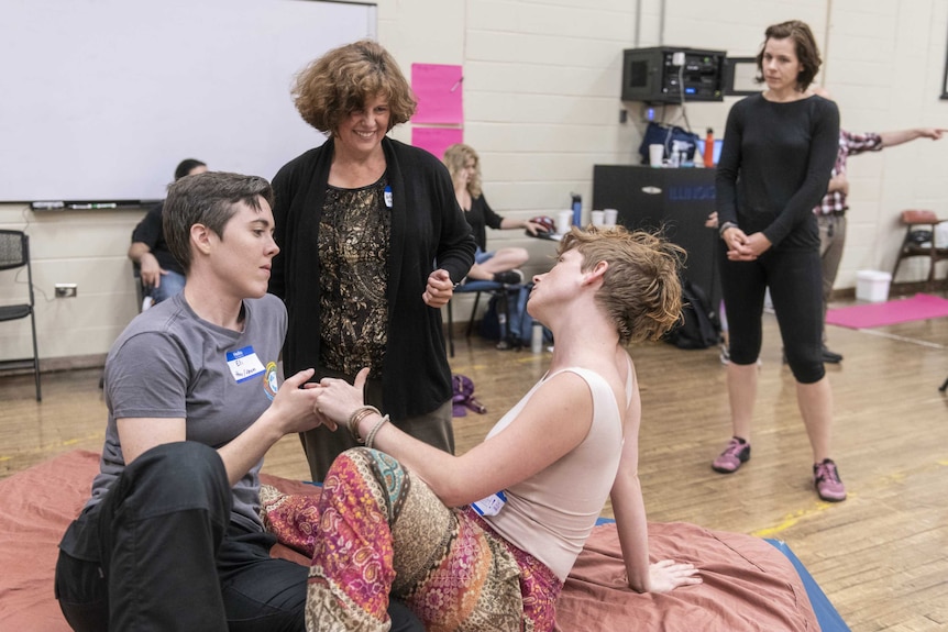 Two actors hold each other's hands and look into each other's eyes while a woman looks on, another woman stands in the back