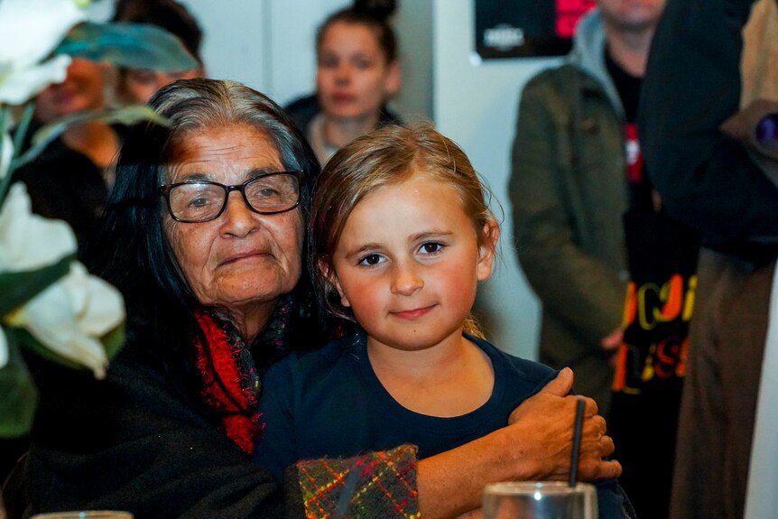 An older woman holds a young child close to her, as they sit in a room surrounded by community.
