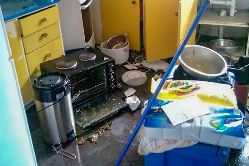 Debris covers a floor in a kitchen with yellow cupboards and blue benchtops. 