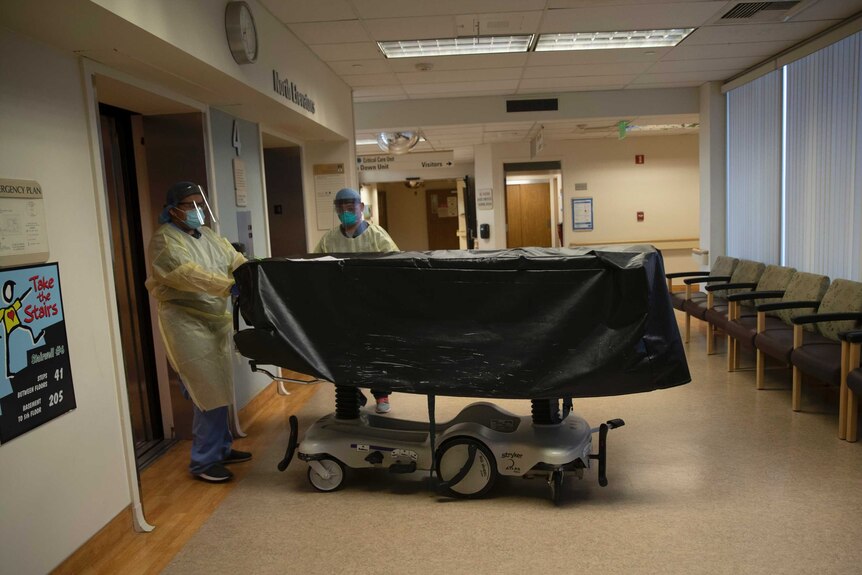 Two hospital workers wearing masks and gowns while pushing a black body bag on wheels