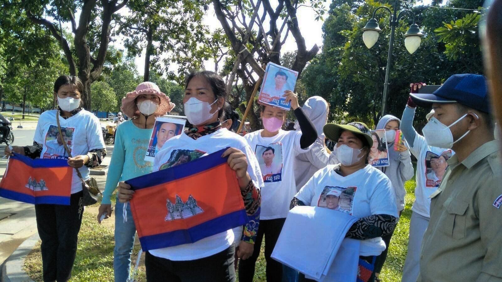 A group of women, many wearing face masks, stand with flags and shirts with men's faces, in a park