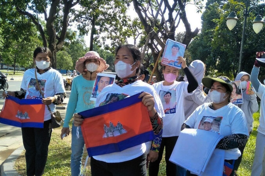 A group of women, many wearing face masks, stand with flags and shirts with men's faces, in a park