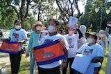 A group of women, many wearing face masks, stand with flags and shirts with men's faces, in a park